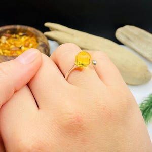 May include: A silver ring featuring a large, round, amber-colored gemstone. The ring has a simple, open design with a small silver ball on one side. The gemstone is a vibrant yellow-orange color.