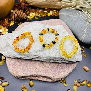 May include: Three amber bracelets with varying shades of yellow and brown beads are displayed on a light gray stone. The bracelets are arranged on a larger, reddish-brown stone, with loose amber pieces scattered around. The background includes natural elements like a wooden bowl and pine cones.