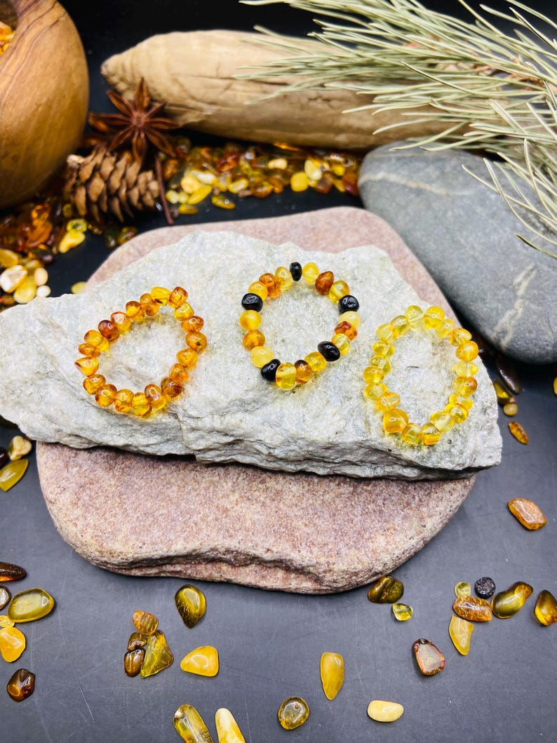 May include: Three amber bracelets with varying shades of yellow and brown beads are displayed on a light gray stone. The bracelets are arranged on a larger, reddish-brown stone, with loose amber pieces scattered around. The background includes natural elements like a wooden bowl and pine cones.