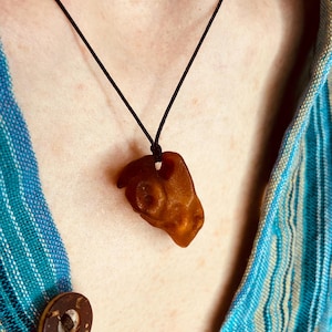 Könnte beinhalten: Eine Halskette mit einem Anhänger aus natürlichem Bernstein. Der Bernstein hat eine warme, goldbraune Farbe und eine unregelmäßige, organische Form. Der Anhänger hängt an einer schwarzen Kordel. Die Person trägt ein blau-weiß gestreiftes Hemd.