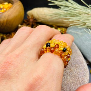 May include: A beaded ring made of amber-colored beads, with a few black beads. The ring is on a person's finger. The ring is a mix of yellow and brown tones. The background includes a wooden bowl and a stone.