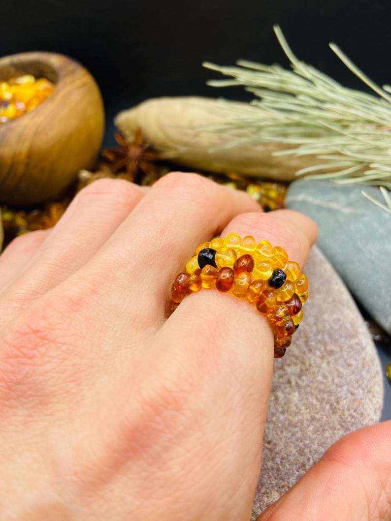 May include: A beaded ring made of amber-colored beads, with a few black beads. The ring is on a person's finger. The ring is a mix of yellow and brown tones. The background includes a wooden bowl and a stone.