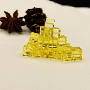 Può includere: Una piramide di piccoli cubi gialli traslucidi. I cubi sono di dimensioni uniformi e sembrano essere fatti di un materiale trasparente. Sullo sfondo, ci sono un anice stellato e una pigna. I cubi misurano circa 1 cm.