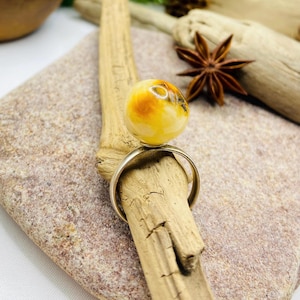 May include: A silver ring featuring a large, round amber stone with yellow and brown hues. The ring is displayed on a piece of driftwood, set against a neutral background with natural elements.
