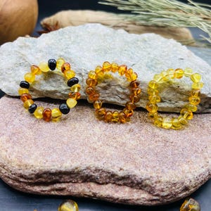 May include: Three rings made of amber beads in varying shades of yellow, orange, and brown. One ring features black beads. The rings are displayed on a gray and brown stone. The amber beads are likely measured in millimeters.