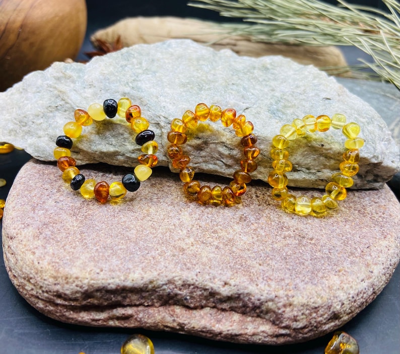 May include: Three amber rings, each composed of small, rounded amber beads. The rings vary in color, with shades of yellow, orange, and dark brown. One ring has black beads interspersed. The rings are displayed on a gray and brown stone surface.