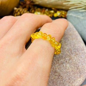 May include: A close-up of a yellow amber ring. The ring is made of small, rounded amber beads strung together. The ring is on a finger, with a natural stone in the background. The amber beads have a translucent quality.