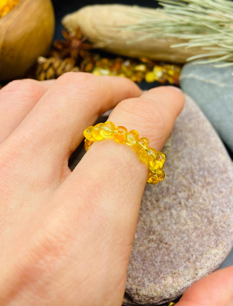 May include: A close-up of a yellow amber ring. The ring is made of small, rounded amber beads strung together. The ring is on a finger, with a natural stone in the background. The amber beads have a translucent quality.