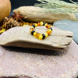 May include: A beaded ring made of amber stones in shades of yellow, orange, and black. The ring is displayed on a piece of driftwood, with a natural stone background. The ring is a unique piece of jewelry.