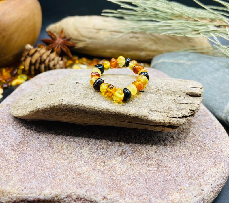 May include: A beaded ring made of amber stones in shades of yellow, orange, and black. The ring is displayed on a piece of driftwood, with a natural stone background. The ring is a unique piece of jewelry.