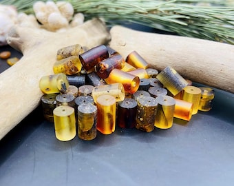 May include: A collection of amber beads in various shades of yellow, brown, and black. The beads are cylindrical and have a smooth, polished finish. They are arranged in a pile on a wooden surface.