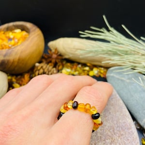 May include: A ring made of amber beads in shades of yellow, orange, and black. The ring is worn on a finger, showcasing the varying colors and textures of the amber stones. The ring is a unique piece of jewelry.