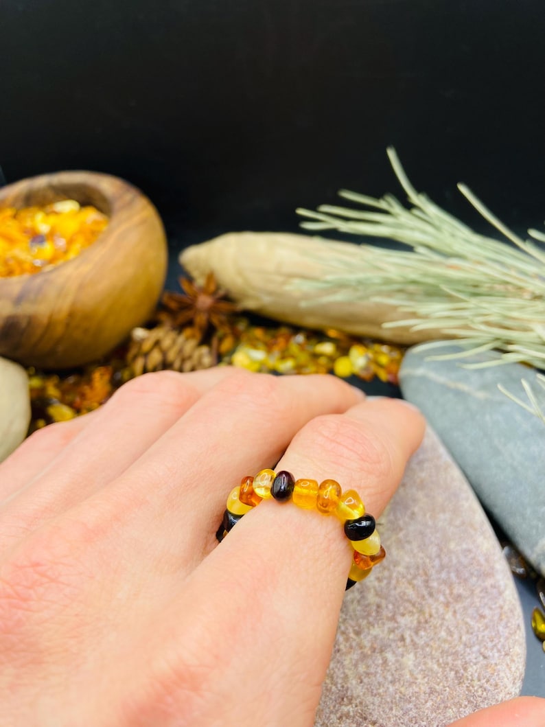 May include: A ring made of amber beads in shades of yellow, orange, and black. The ring is worn on a finger, showcasing the varying colors and textures of the amber stones. The ring is a unique piece of jewelry.