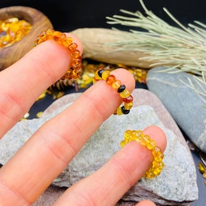 May include: Three amber beaded rings are displayed on fingers. The rings are made of small, rounded amber beads in shades of yellow, orange, and brown. One ring has black beads. The rings are on a gray stone surface.