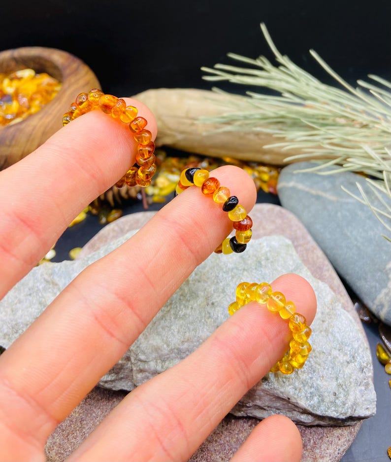 May include: Three amber beaded rings are displayed on fingers. The rings are made of small, rounded amber beads in shades of yellow, orange, and brown. One ring has black beads. The rings are on a gray stone surface.
