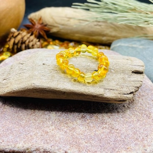 May include: A golden amber beaded ring. The ring is composed of small, round, translucent beads. The ring is displayed on a piece of driftwood, with a natural stone background. Other natural elements are in the background.