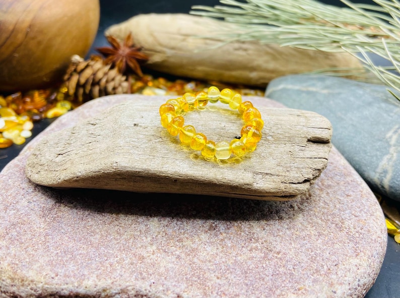 May include: A golden amber beaded ring. The ring is composed of small, round, translucent beads. The ring is displayed on a piece of driftwood, with a natural stone background. Other natural elements are in the background.