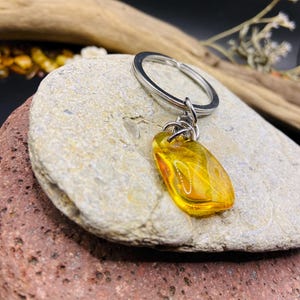 Può includere: Un ciondolo portachiavi rettangolare color ambra con un portachiavi color argento. Il ciondolo ha un aspetto lucido e traslucido, che mostra motivi interni. Il portachiavi è esposto su una pietra di colore chiaro, con uno sfondo scuro.