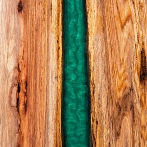 May include: A wooden board with a green epoxy resin inlay. The wood has a natural grain pattern and the resin has a swirling pattern.