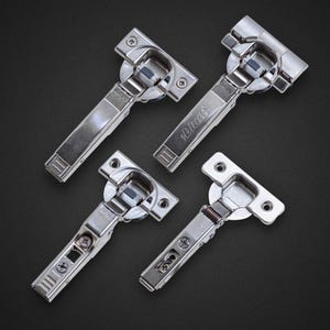 May include: Four shiny, silver cabinet hinges arranged on a dark background. Each hinge has a rectangular body with a curved section and mounting plate. The brand name "blum" is visible on the hinge.