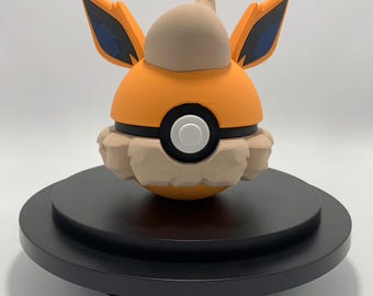 May include: A 3D printed figurine of a Pokemon character, Vaporeon, in its Poké Ball form. The figurine is orange, brown, and blue, and is sitting on a black circular base.