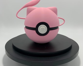 Könnte beinhalten: Eine rosa, kugelförmige Skulptur, die einer Zeichentrickfigur ähnelt, mit einem schwarzen Band und einem weißen Kreisdetail. Sie hat ohrartige Vorsprünge und ein gebogenes, antennenartiges Element. Die Skulptur wird auf einer schwarzen, mehrstufigen Plattform ausgestellt.