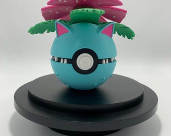 May include: A blue and green Pokemon figure with a pink flower on top. The figure is standing on a black circular base.
