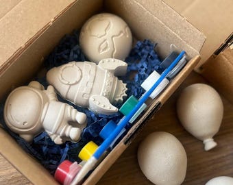 May include: A craft kit in a cardboard box containing a rocket, astronaut, and planet. The kit includes paint pots, a paintbrush, and balloon-shaped objects. The items are beige and sit on blue paper filler.