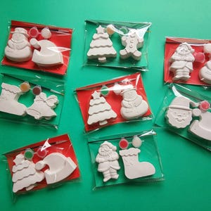 Puede incluir: Un conjunto de kits de manualidades navideñas para pintar. Cada kit incluye una figura de yeso blanco, como un muñeco de nieve, Papá Noel o un árbol de Navidad, con pintura y un pincel, todo empaquetado en una bolsa de plástico transparente con respaldo rojo.