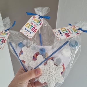 Puede incluir: Una bolsa de celofán transparente contiene un kit de manualidades con forma de copo de nieve con botes de pintura, un pincel y una etiqueta que dice "THANKS for making your BIRTHAY EXTRA special". El kit está atado con una cinta azul.