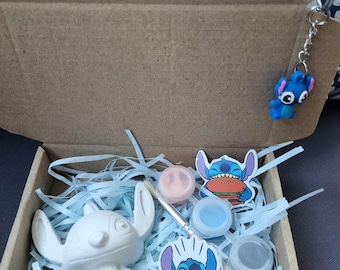 May include: A Stitch-themed paint-your-own craft kit in an open cardboard box. The kit contains a white Stitch figurine, paint pots, a paintbrush, and Stitch stickers. A blue Stitch keyring hangs from the top. The box is filled with blue paper shreds.
