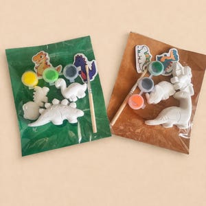 Puede incluir: Dos kits de manualidades con figuras de dinosaurios, botes de pintura y pinceles. Un kit está en una bolsa verde, el otro en una bolsa marrón. Las figuras son blancas y los botes de pintura contienen pintura amarilla, verde, naranja y azul.