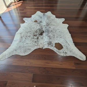 Könnte beinhalten: Ein Kuhfellteppich mit einem natürlichen Muster aus weißen, grauen und braunen Flecken. Der Teppich hat eine unregelmäßige Form und liegt auf einem dunklen Holzboden. Das Fell hat ein weiches, strukturiertes Aussehen.