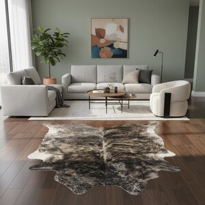 Puede incluir: Salón con una alfombra de piel de vaca con marcas marrones y blancas sobre un suelo de madera oscura. Sofá gris claro, sillón y mesa de centro con objetos decorativos. Una pintura abstracta cuelga de la pared y una planta en maceta añade vegetación.