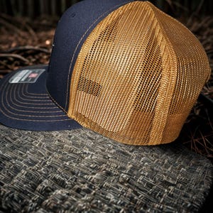 Może przedstawiać: Granatowa czapka typu trucker ze złotą siatką z tyłu. Czapka ma zakrzywiony daszek i strukturalny panel przedni. Szwy są złote, a czapka spoczywa na teksturowanej powierzchni.