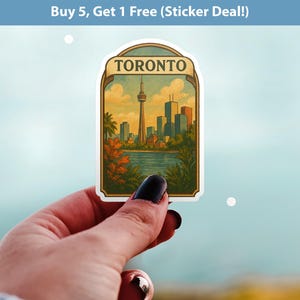 Peut inclure: Un autocollant rectangulaire avec une illustration de style vintage de l'horizon de Toronto, comprenant la Tour CN et d'autres bâtiments. L'autocollant a une bordure crème et le mot "TORONTO" en haut. L'arrière-plan est une image floue d'une main tenant l'autocollant.