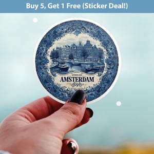 Puede incluir: Una pegatina redonda con una ilustración azul y blanca de Ámsterdam, Países Bajos. El diseño incluye edificios, canales y el nombre de la ciudad. La pegatina se sostiene sobre un fondo borroso.