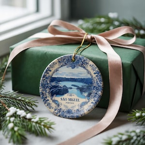 May include: A round ceramic ornament with a blue and white floral design and a landscape scene of a lake and mountains. The ornament is hanging in front of a wrapped gift box with a pink ribbon. The text "SÃO MIGUEL PORTUGAL" is visible.