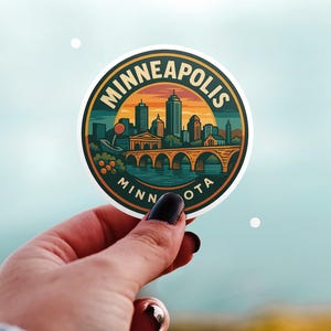 May include: A round sticker featuring a Minneapolis, Minnesota, skyline with a bridge over water. The sticker has a sunset background and the words "MINNEAPOLIS MINN OTA" in a circular design. Held by a hand.