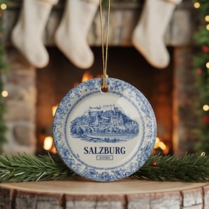 May include: A circular ceramic ornament with a blue and white design depicting Salzburg, Austria. The ornament has a floral pattern around the edge and the word "SALZBURG" is printed in the centre. A gold string is attached for hanging.