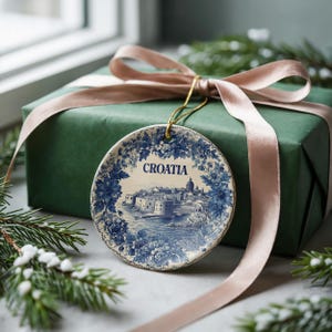 Puede incluir: Adorno redondo de cerámica con una ilustración azul y blanca de una ciudad costera y la palabra "CROATIA". Cuelga frente a una caja de regalo verde atada con una cinta rosa, con vegetación festiva.