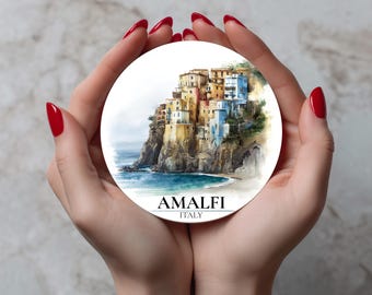 May include: A round coaster featuring a watercolor painting of Amalfi, Italy. The artwork depicts colorful buildings on a cliffside overlooking the sea. The text "AMALFI ITALY" is printed at the bottom.