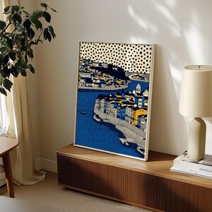K&ouml;nnte beinhalten: Gerahmter Kunstdruck, der eine K&uuml;stenstadt mit blauem Wasser und Geb&auml;uden in Gelb, Blau und Wei&szlig; zeigt. Der Himmel hat ein Muster aus schwarzen Punkten. Das Kunstwerk wird auf einem Holzschrank ausgestellt.