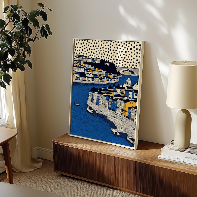 K&ouml;nnte beinhalten: Gerahmter Kunstdruck, der eine K&uuml;stenstadt mit blauem Wasser und Geb&auml;uden in Gelb, Blau und Wei&szlig; zeigt. Der Himmel hat ein Muster aus schwarzen Punkten. Das Kunstwerk wird auf einem Holzschrank ausgestellt.