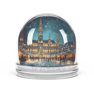 May include: A snow globe with a silver base and a clear glass dome. The interior features a detailed illustration of a building, a Christmas tree, and people in a winter scene. The background is a dark blue with falling snow.