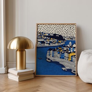 K&ouml;nnte beinhalten: Eine goldfarbene, pilzf&ouml;rmige Tischlampe steht auf einem Stapel B&uuml;cher neben einem gerahmten Kunstdruck. Der Druck zeigt eine K&uuml;stenstadt mit blauem Wasser, Geb&auml;uden in Blau, Gelb und Wei&szlig; und einem gepunkteten Himmel.