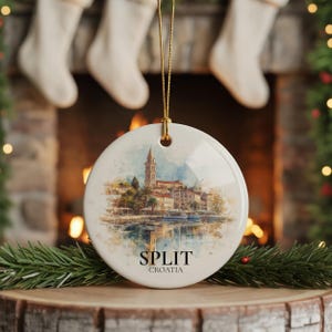 Puede incluir: Una bola de Navidad redonda de cerámica blanca con una pintura de acuarela de Split, Croacia. La imagen muestra una ciudad costera con una alta torre de iglesia, barcos y el texto "SPLIT CROACIA" en negro. Una cuerda dorada está adjunta.
