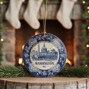 May include: A round, blue and white ceramic Christmas ornament featuring a detailed illustration of the Washington D.C. Capitol building and the text "WASHINGTON D.C.". The ornament has a floral border and a gold string for hanging.