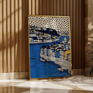 K&ouml;nnte beinhalten: Gerahmter Kunstdruck mit einer detaillierten Illustration einer K&uuml;stenstadt mit blauem Wasser und Geb&auml;uden in Wei&szlig;, Gelb und Blau. Das Kunstwerk hat einen goldenen Rahmen und einen gepunkteten Hintergrund. Der Druck wird vor einer Holzwand ausgestellt.