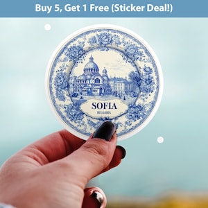 May include: A round sticker featuring a blue and white illustration of a building in Sofia, Bulgaria, with floral accents. The sticker has the word "SOFIA" and "BULGARIA" printed on it. The sticker is held up against a blurred background.